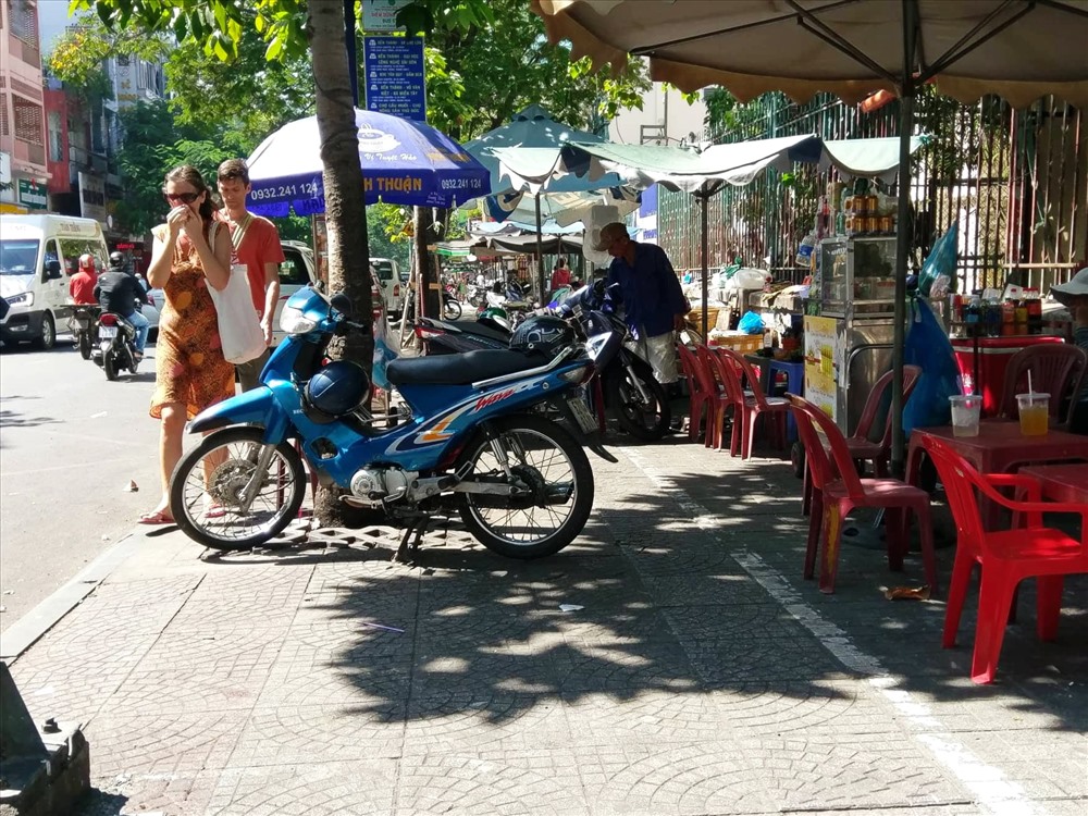 Vỉa hè đường Nguyễn Thái Bình bị chiếm dụng để bàn ghế, xe máy trông lụp xụp và nhếch nhác. Trong ảnh người đi bộ bị chắn lối đi nên phải đi ra sát lề đường.
