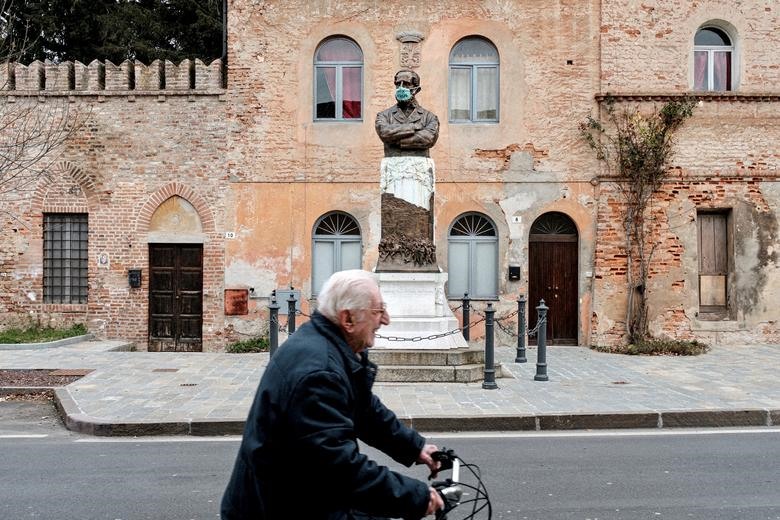 Một người đàn ông lớn tuổi không đeo khẩu trang, đạp xe ngang qua một bức tượng đeo khẩu trang ở San Fiorano, ngày 28.2. Ảnh: Reuters