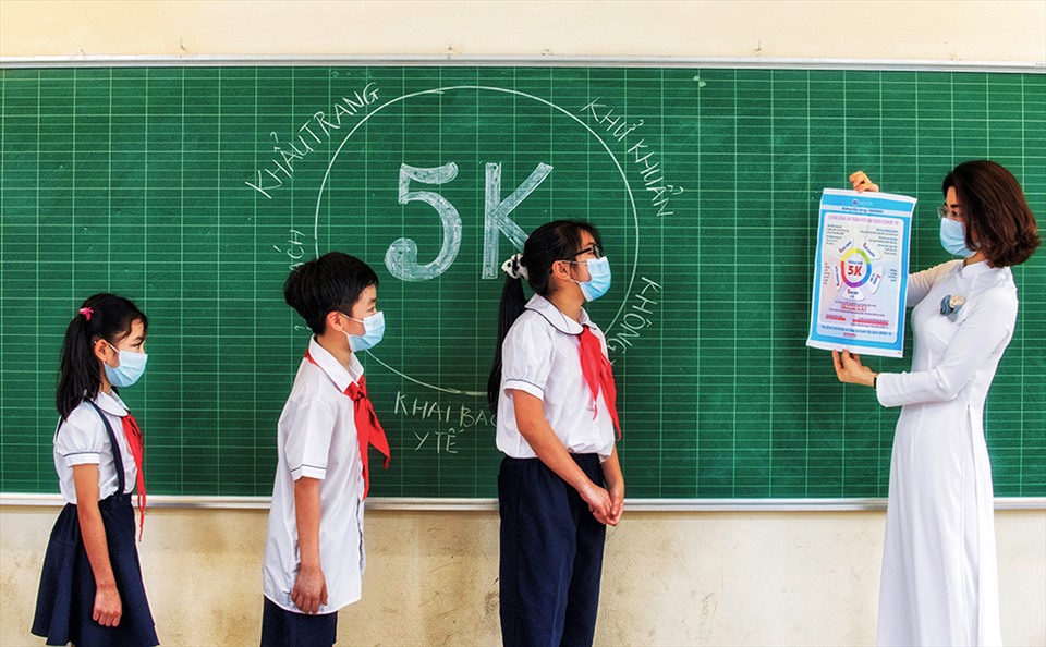 Bài học mới. Cô giáo Ngô Thị Mai Trang trong tà áo dài đang hướng dẫn các em học sinh trường tiểu học Gia Thụy, Hà Nội, Việt Nam 5 biện pháp phòng chống đại dịch Covid-19 Khẩu trang – Khử khuẩn – Khoảng cách – Không tập trung – Khai báo y tế” (5 K) ngày 24.4.2021. Chỉ một thời gian ngắn sau khi bức ảnh này được chụp là tất cả học sinh ở Hà Nội phải ở nhà học online cả một thời gian dài khi Hà Nội bùng phát đợt dịch lần thứ 4.