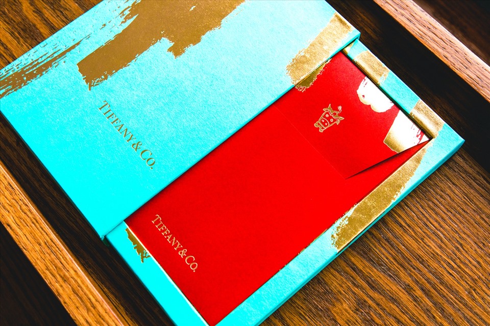 Tiffany & Co. vẫn duy trì màu sắc xanh lam đặc trưng của mình trên hộp phong bì nhưng kết hợp vàng và đỏ ở dạng dập nét cọ và túi giấy đỏ.