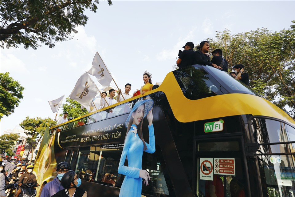 Đứng trên xe buýt hai tầng được trang trí cực sang trọng, Hoa hậu Thùy Tiên diện áo dài vàng, đội vương miện Miss Grand International đứng trên xe buýt 2 tầng. Chưa dừng lại ở đó, xe buýt chở Hoa hậu Thùy Tiên còn được “hộ tống” bởi 4 xe jeep nhỏ. Trên thân của chiếc xe jeep đầu tiên là phiên bản chiếc vương miện mang ý nghĩa to lớn của Việt Nam trên đấu trường nhan sắc quốc tế.