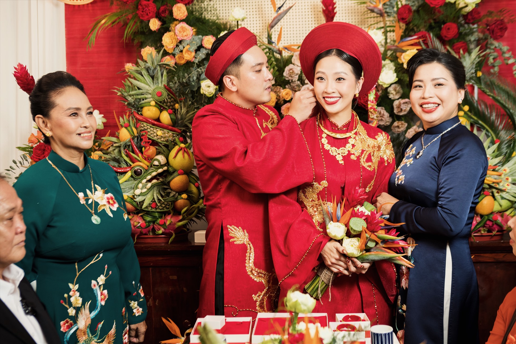 Chiếc áo dài mà nữ MC lựa chọn xuất hiện trong lễ gia tiên cũng mang ý nghĩa đặc biệt và do đích thân NTK Huỳnh Bảo Toàn may tặng riêng cho 2 vợ chồng. Vẫn là hình ảnh áo dài được thêu Long – Phung quen thuộc với sắc đỏ tượng trưng cho điềm may mắn, biểu tượng hạnh phúc, nhưng trang phục của “cô dâu” Hà Trinh lại lấy cảm hứng từ tà áo dài cung đình của Nam Phương Hoàng Hậu, có áo choàng khoác ngoài và đội mấn.