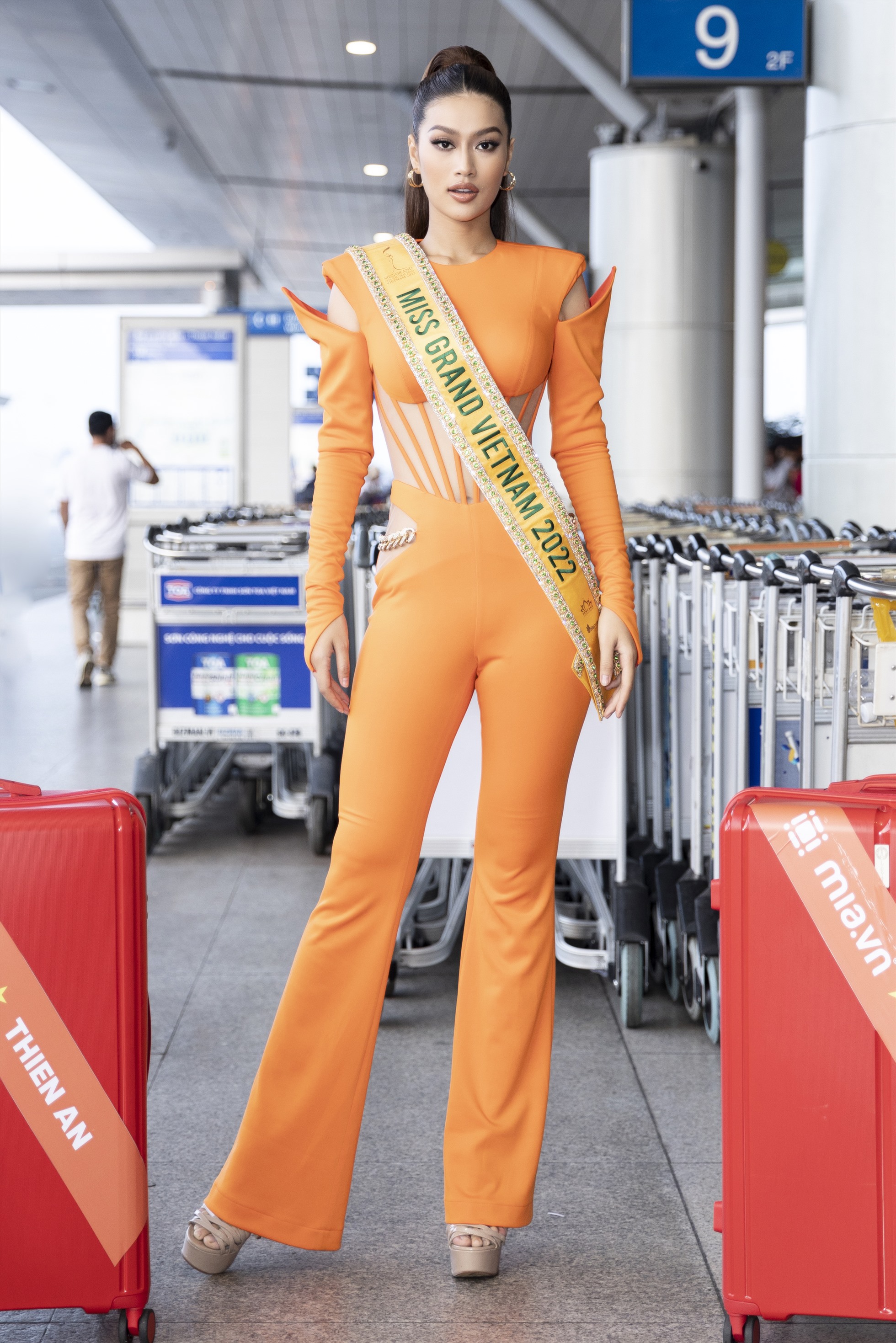 Mặc dù vậy nhưng 6 giờ sáng nay tại sân bay, Thiên  Ân vẫn xuất hiện với vẻ ngoài rạng rỡ tươi tắn. Cô chọn cho mình thiết kế tông cam nổi bật đến từ NTK Tăng Thành Công. Cùng với đó mang theo chiếc sash đại diện “Miss Grand Vietnam” và lá cờ Việt Nam đầy tự hào. Ảnh: NVCC.