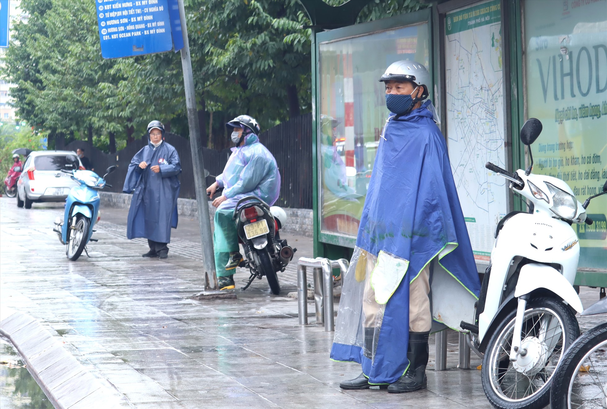Người đàn ông làm nghề xe ôm phải đứng nép vào nhà chờ xe buýt để tránh mưa.