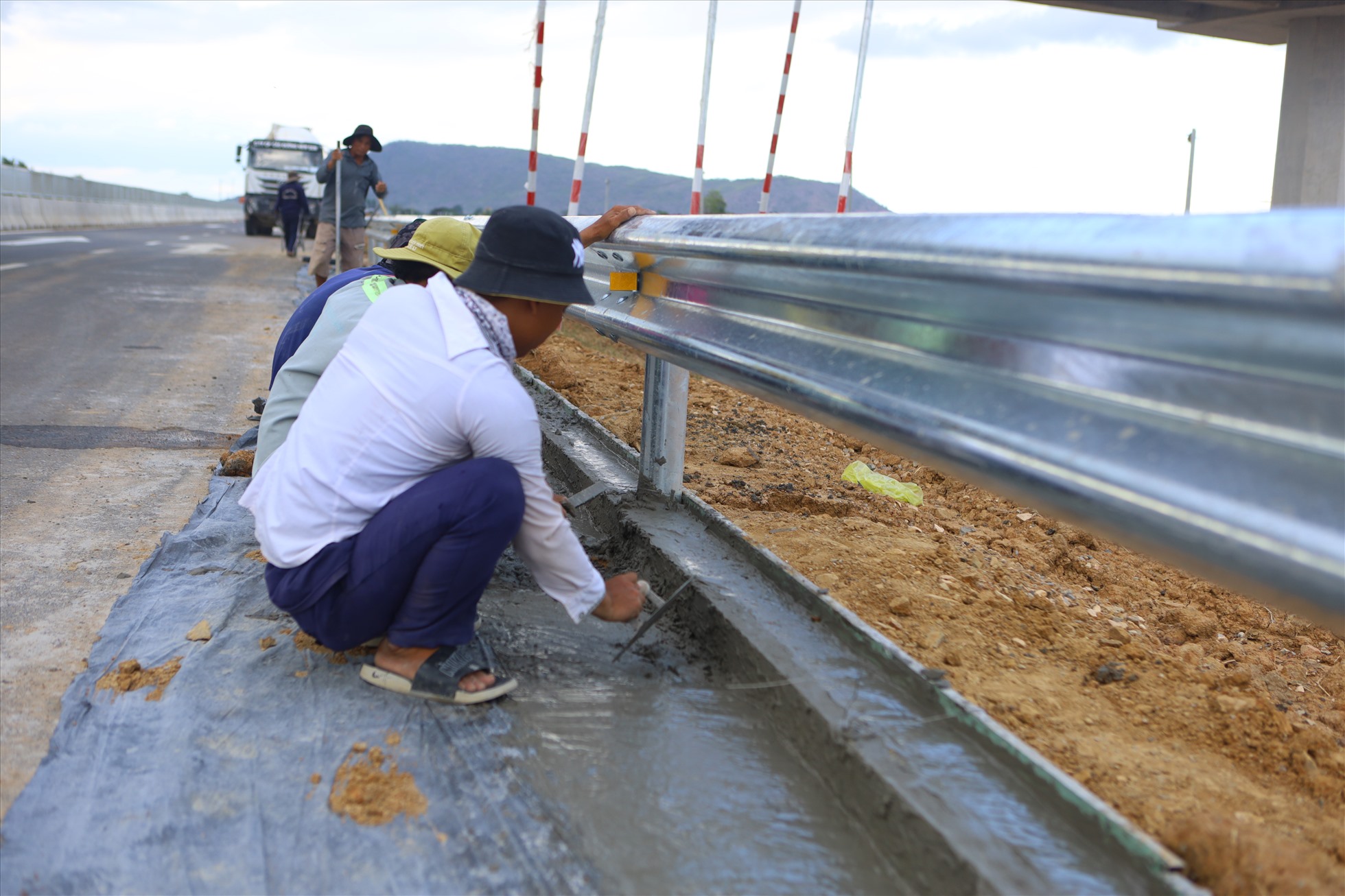 Công nhân đang đổ bê tông gia cố phần chân hộ lan sát mép đường nhựa. “Những ngày này ai nấy cũng khẩn trương thi công theo yêu cầu của đơn vị thi công nên anh em chúng tôi cũng cố gắng để kịp tiến độ”, một công nhân đang thi công chia sẻ.