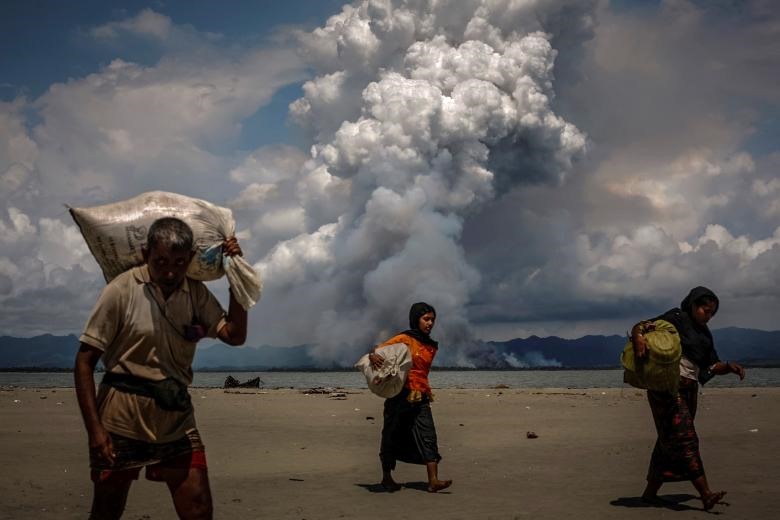 Trong khi những người tị nạn Rohingya đang đi bộ trên bờ biển sau khi đã vượt qua biên giới Bangladesh – Myanmar bằng thuyền qua vịnh Bengal, ở Shah Porir Dwip, Bangladesh; thì phía biên giới Myanmar, những đám khói lớn vẫn đang bốc lên nhiều hơn, ngày 11/09/2017. (Ảnh: Reuters)