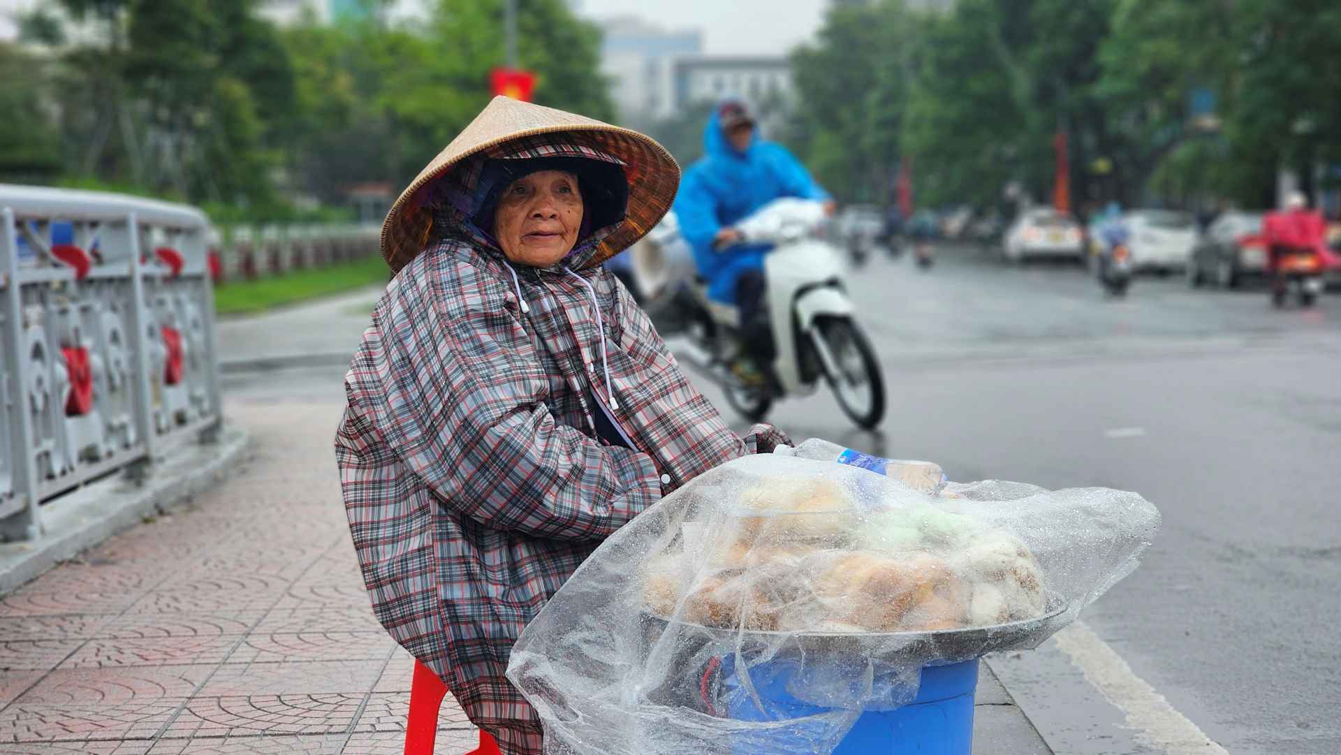Bà Lê Thị Lệ (60 tuổi) quấn mình trong chiếc áo mưa, ngồi co ro giữa trời để bán bánh ngọt ở cầu Kho Rèn.  