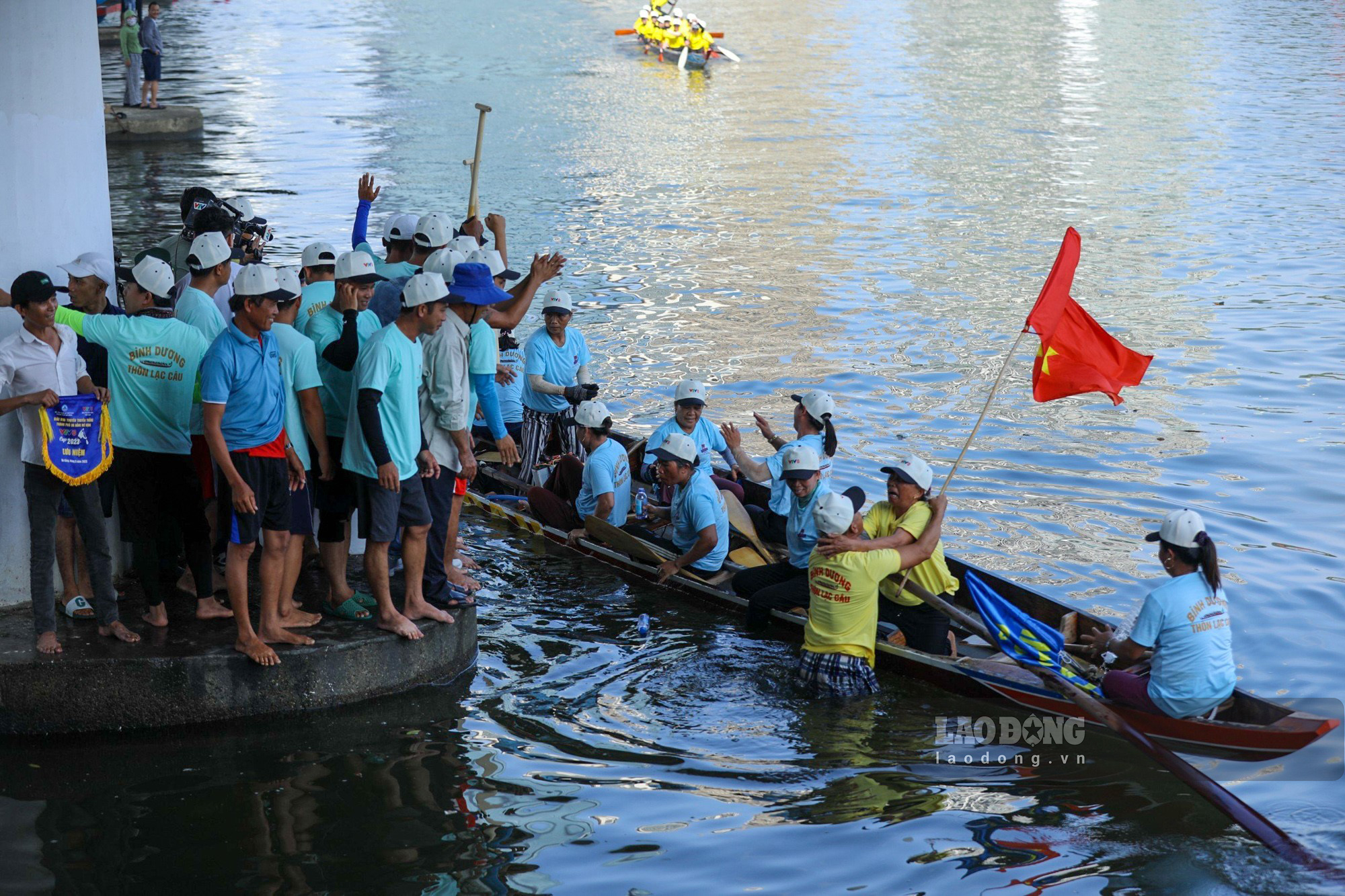 Một đội đua vừa hoàn thành chặn đua với thứ hạng đứng đầu, người cổ vũ ào ra ăn mừng khi thuyền cập bờ.