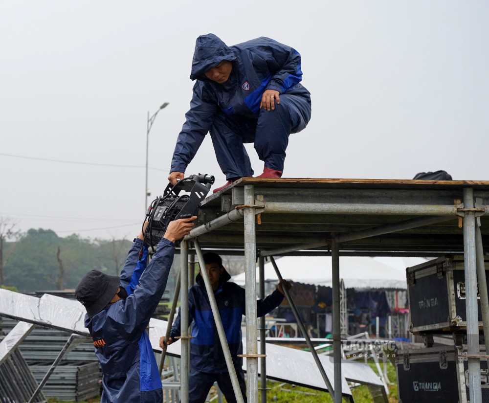 Dưới trời mưa lạnh, công tác thi công lắp ráp sân khấu diễn ra khá khó khăn. Ngoài việc trang bị đồ bảo hộ, các công nhân phải mặc thêm áo mưa. 