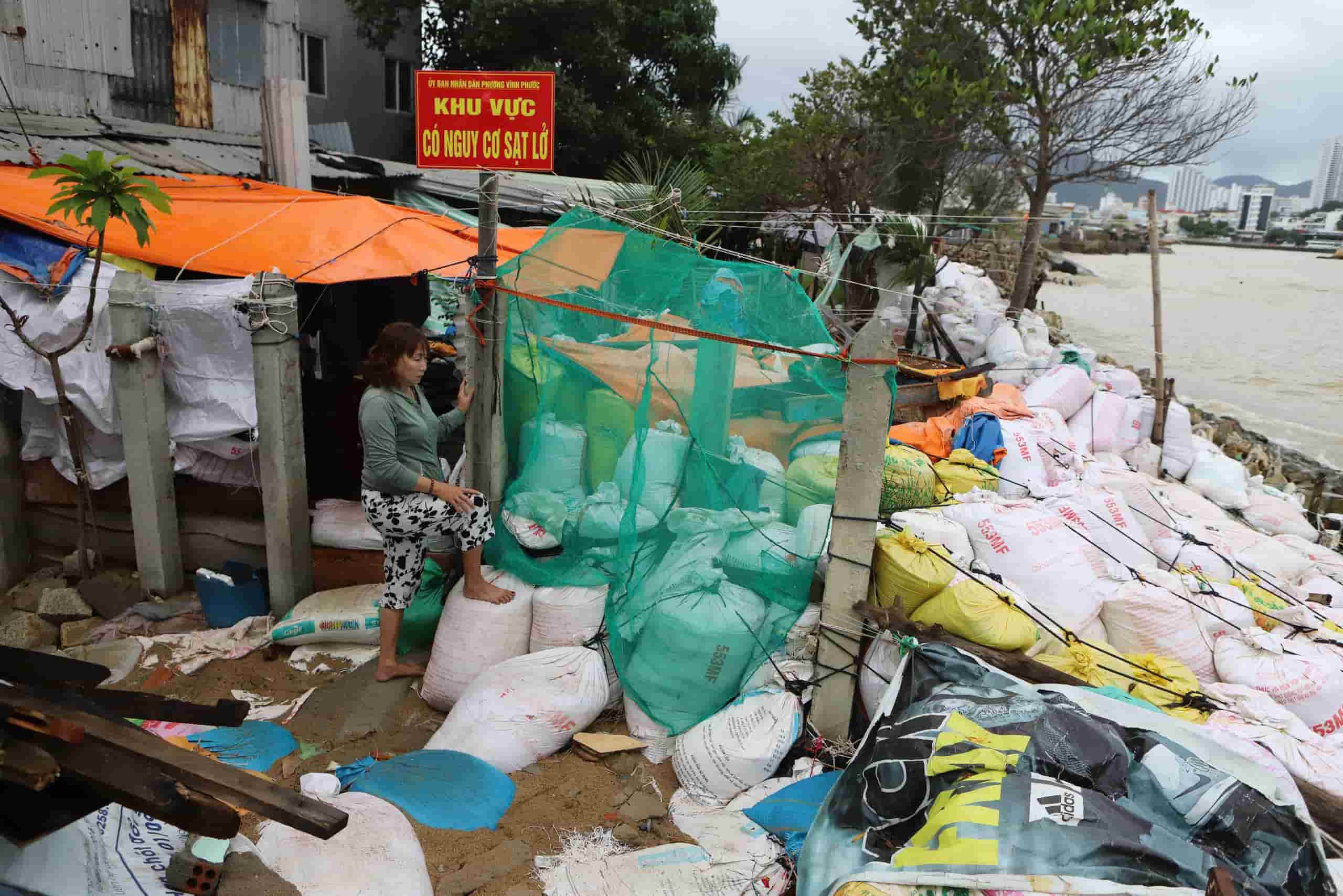 nhà bà Nguyễn Thị Bé (51 tuổi) cũng bị sóng đánh. Thời gian qua, bà Bé phải dùng rọ đá, thanh gỗ, bao cát để gia cố nhà.  Người phụ nữ chia sẻ chồng bà làm nghề biển nên vắng nhà liên tục. Vì vậy, mỗi mùa biển động, bà cứ nơm nớp lo sợ.  Hôm 27/12, sóng đánh rất mạnh. Bà Bé tranh thủ lúc ngớt mưa ra kiểm tra “hệ thống phòng thủ” chắn sóng.  “Vợ chồng tôi làm được bao nhiêu tiền đều phải trích ra mua đá, bao tải đắp bờ kè vì sợ sóng đánh bay mất nhà” - người phụ nữ than thở.