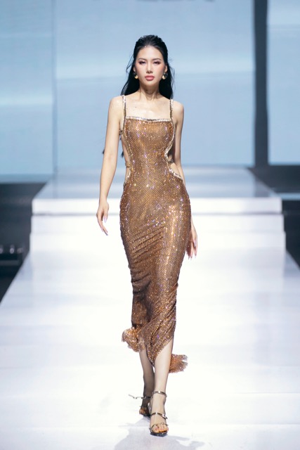 Cũng trong đêm thứ hai của Vietnam International Fashion Week 2024, Bùi Quỳnh Hoa chinh phục trái tim khán giả ngay từ khoảnh khắc đầu tiên sải bước với vai trò First face cho BST Shades Of Midnight của CEM By Lê Minh Ngọc. Khoác lên mình thiết kế vàng được làm từ chất liệu sequin ánh kim lấp lánh, nàng hậu như hóa thân thành một quý cô hào nhoáng trong bữa tiệc. Chiếc váy hai dây hở lưng cùng phần thân váy bó sát giúp người đẹp khoe vóc dáng cực quyến rũ.  