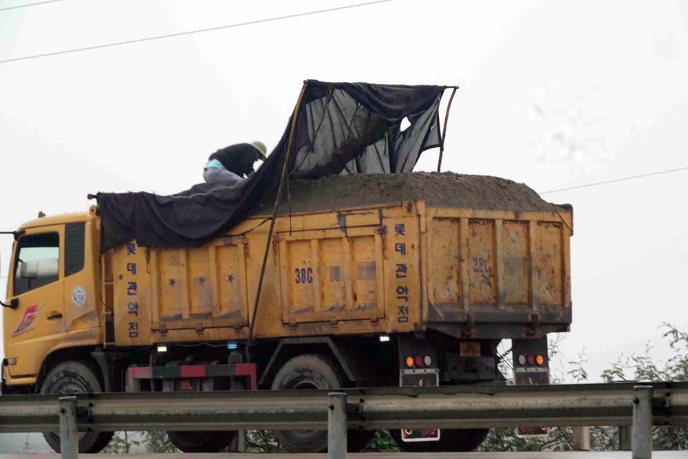 Một xe tải chở cát chất quá thành thùng từ bãi tập kết cát ở phường Bắc Hồng Lĩnh vừa di chuyển ra Quốc lộ 1. Tài xế đang kiểm tra, đậy lại bạt trước khi tăng tốc lưu thông. Ảnh: Trần Tuấn