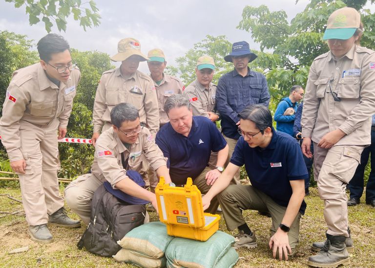 El viceministro de Relaciones Exteriores de Vietnam, Do Hoang Viet (quien esta a la derecha de la foto) y el embajador de los Estados Unidos en Vietnam, Marc Knapper (quien esta sentado en el centro de la foto) presionan para detonar las bombas. Imagen de la pagina web de la organizacion