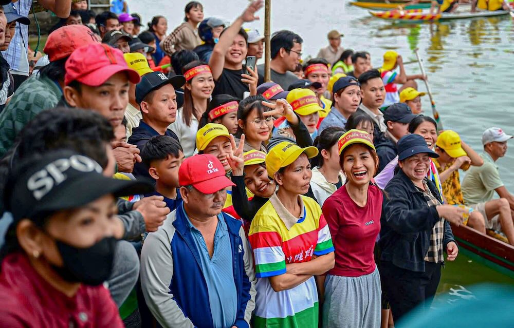 Trên bờ, cờ phướn tung bay trong nắng Xuân. Dòng người cổ vũ tạo nên bức tranh sinh động, đậm chất lễ hội làng quê ven sông.