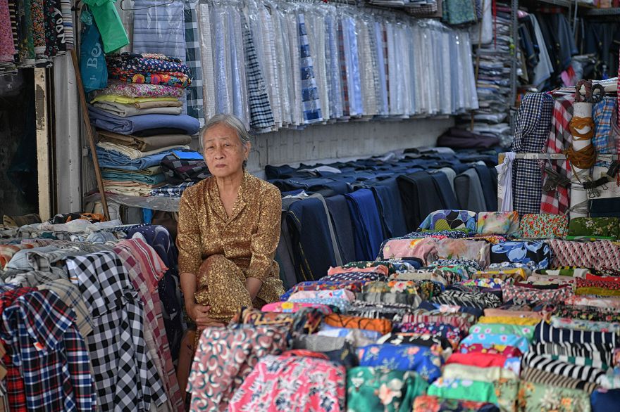 Một bà cụ ngồi trông quầy giữa các chồng vải xếp gọn, gợi lên hình ảnh gắn bó lâu năm của những người bán với khu chợ truyền thống. Ảnh: Việt Văn