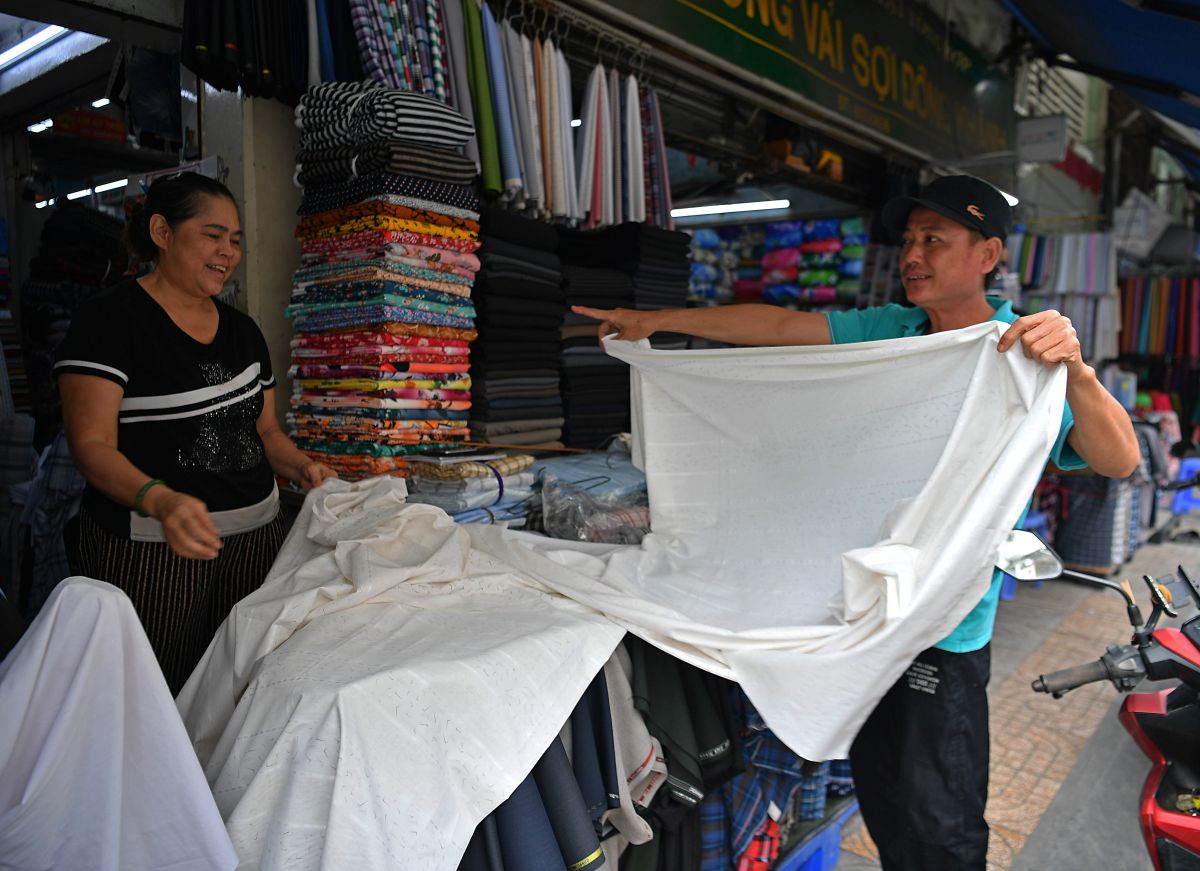 Tiểu thương tại chợ vải giới thiệu chất liệu cho khách ngay trước gian hàng, phản ánh hoạt động mua bán sôi động và tính tương tác trực tiếp đặc trưng của chợ truyền thống. Ảnh: Việt Văn