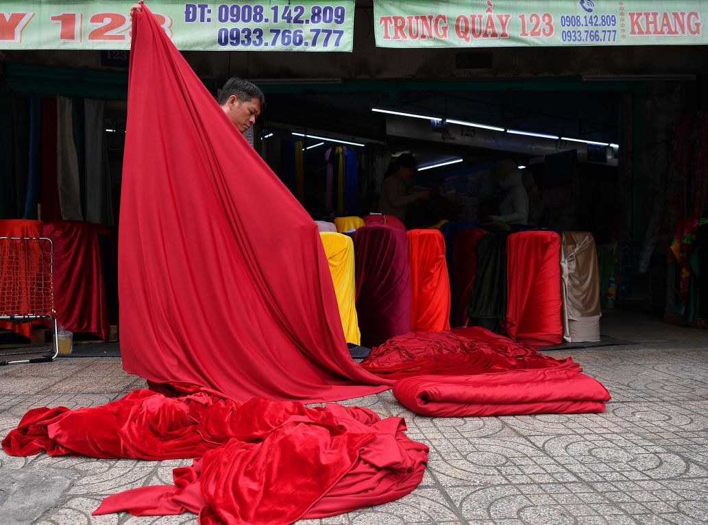 A red cloth stretched from the seller's hand, spilling onto the sidewalk like a soft flow of color, standing out in the daily trading rhythm at Soai Kinh Lam fabric market. Photo: Viet Van
