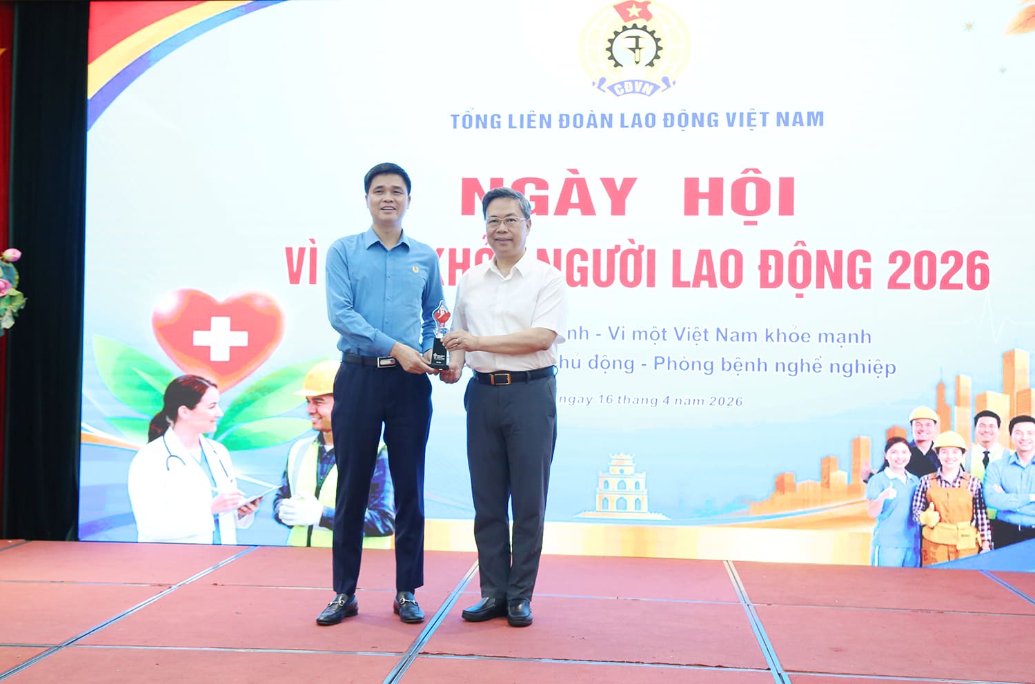 Ông Nguyễn Hà Thanh (áo trắng) - Viện trưởng Viện Huyết học - Truyền máu Trung ương tặng biểu trưng linh vật hiến máu cho đại diện Ban tổ chức chương trình “Ngày hội vì sức khoẻ Người lao động” 2026. Ảnh: Vương Tuấn. 
