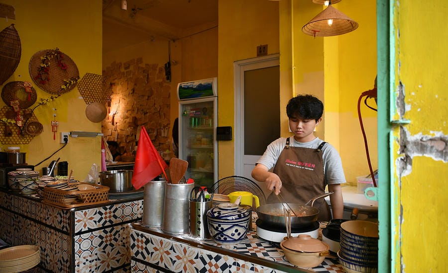 Một nhân viên trẻ đang chế biến món ăn tại quầy bếp được trang trí theo phong cách hoài cổ với gạch bông và nón lá. Ảnh: Việt Văn