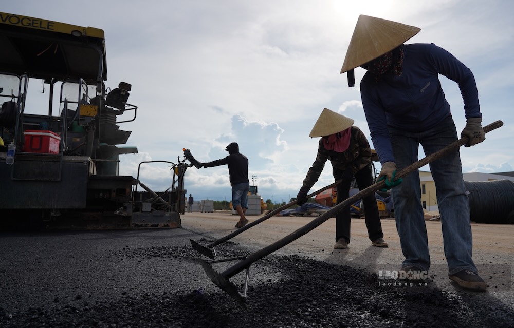 Hai công nhân nam đi sát hai bên máy rải để căn chỉnh hướng thảm, bảo đảm mặt đường được thi công đúng thiết kế và thẳng tuyến. Phía sau, các nữ công nhân sử dụng cào chuyên dụng để san gạt, chỉnh sửa lớp bê tông nhựa nóng, giúp bề mặt đường bằng phẳng trước khi lu lèn. 
