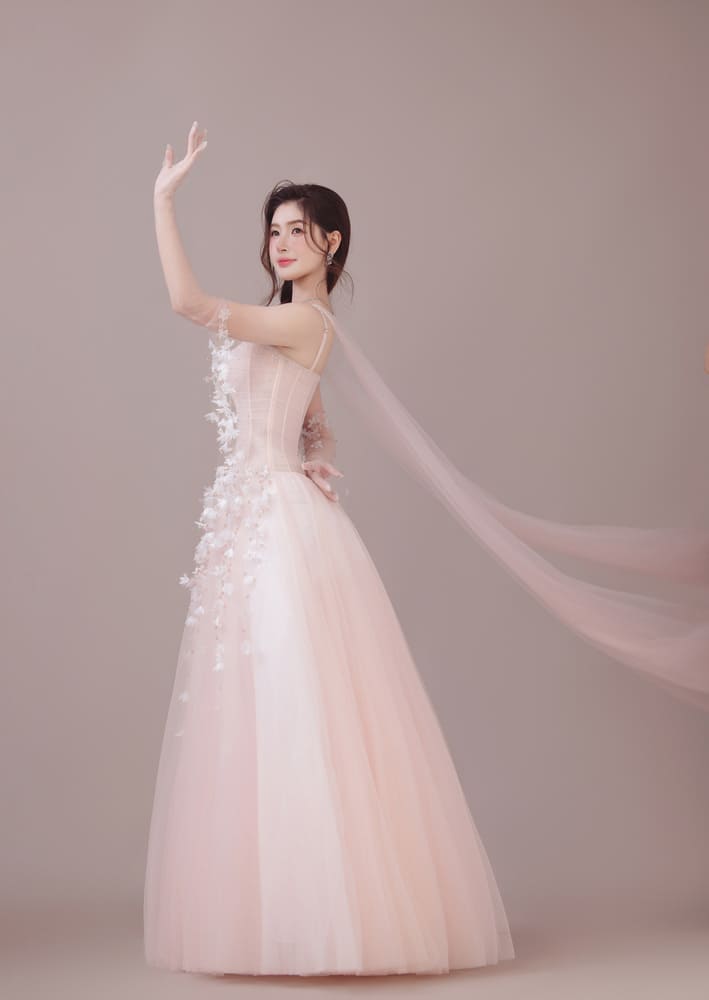 Thu Uyên chọn đầm dạ hội màu hồng phấn trong bộ ảnh mới. Thiết kế ôm phần thân trên với hai dây mảnh, chân váy xòe rộng, phủ nhiều lớp tulle. Những chi tiết hoa 3D được đính dọc thân và chân váy, kết hợp tà voan dài phía sau. Người đẹp dùng găng tay xuyên thấu đính hoa và trang sức tối giản.